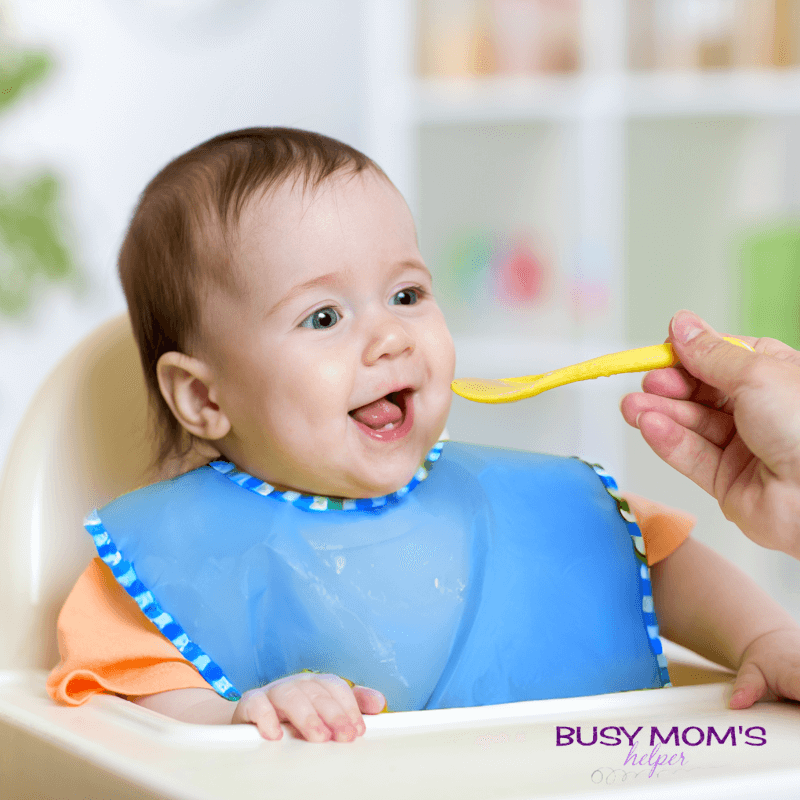 baby eating food from a spoon in a blue bib with a orange shirt.